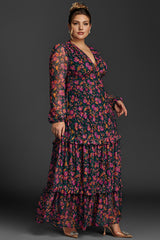 A woman wearing a vibrant, floral maxi dress with long sheer sleeves and tiered ruffles, posing confidently against a dark background.